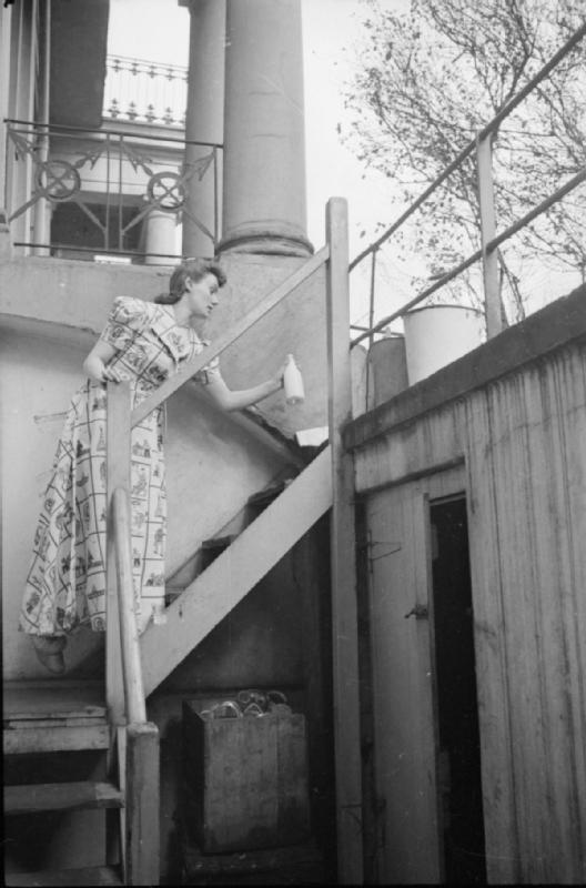 Mrs Olive Day va chercher le lait et le journal posés en haut des escaliers qui emmènent vers son sous sol. Les seaux  contiennent du sable et de l'eau et étaient fournis en cas de bombe incendiaire.
