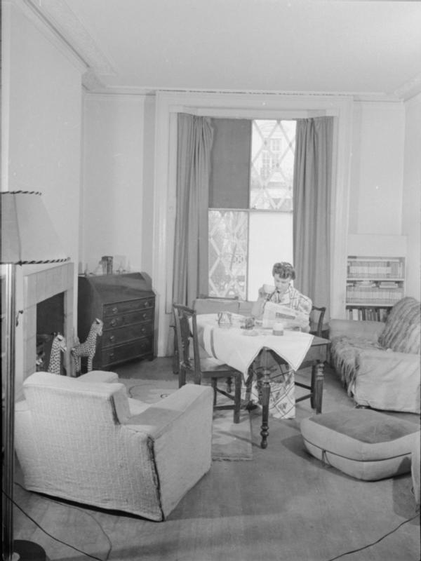 Mrs Olive Day bois son thé en lisant le journal dans son salon, certaines vitres de la fenêtre ont été cassée par une bombe, les autres sont  recouvertes de scotch pour éviter les éclats.